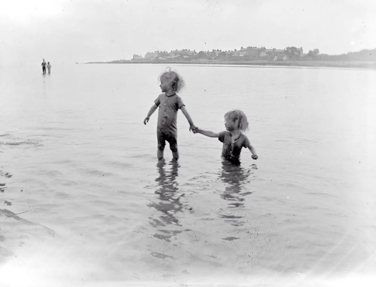"Two little children bathing in seaside two other figures in distance, town visible in background" This…